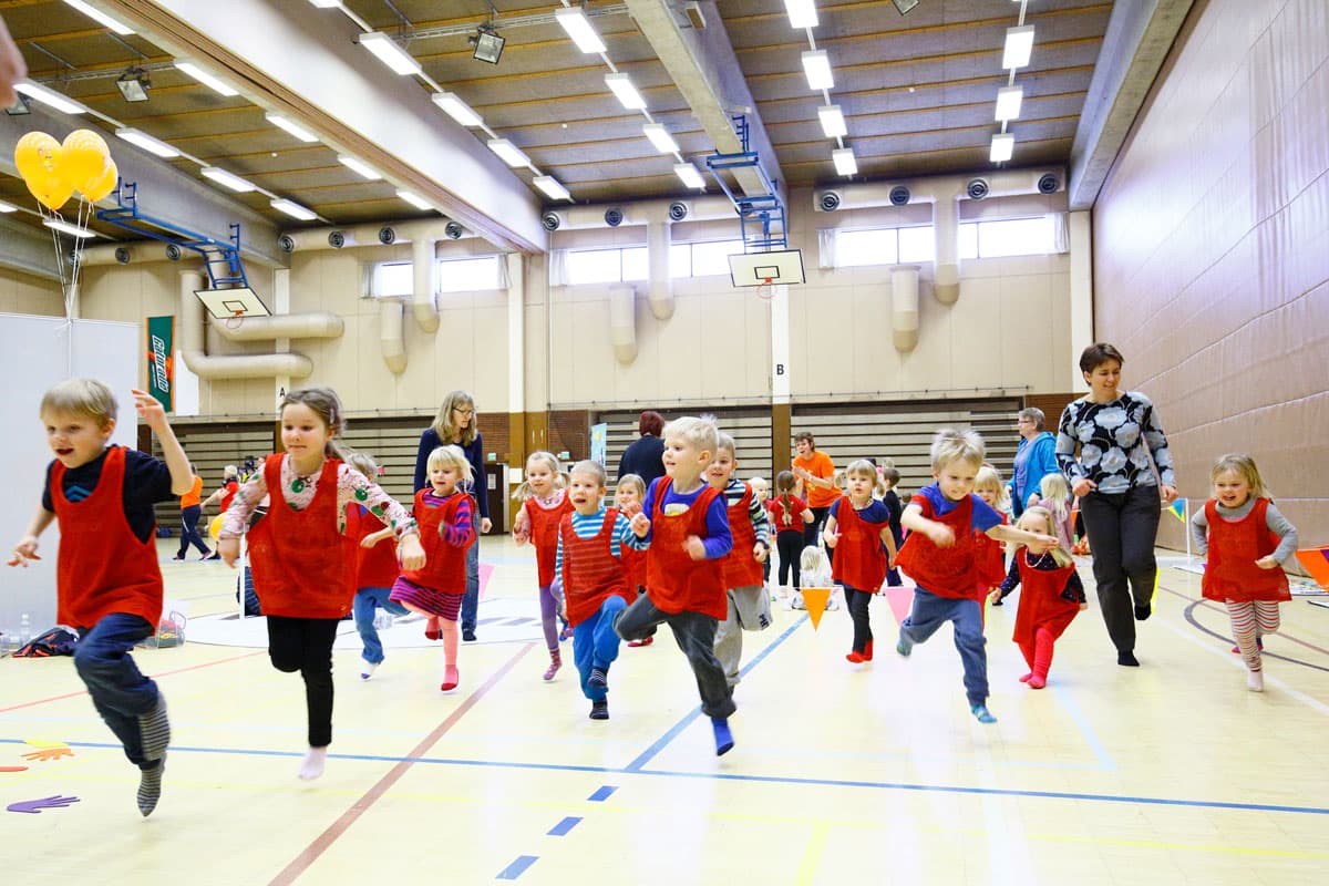 En grupp glada barn i röda västar springer och leker i en gymnastiksal, uppmuntrade av vuxna ledare i bakgrunden.