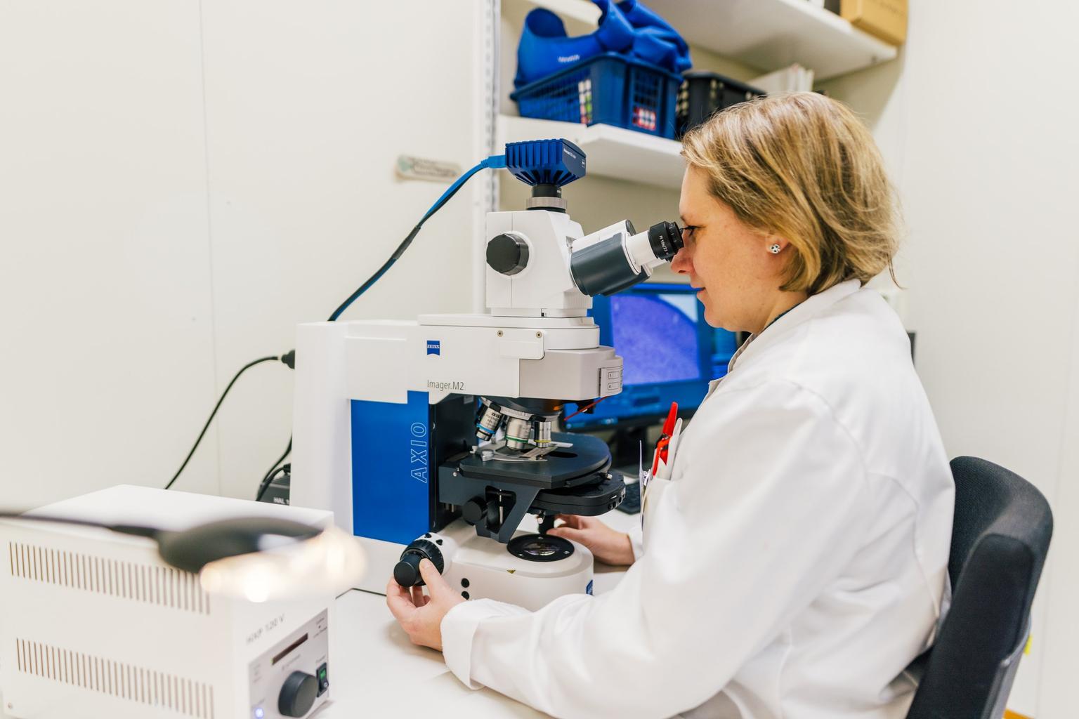A scientist wearing a white lab coat is looking through a microscope in a laboratory. She is adjusting the focus knobs while a computer screen in the background displays a magnified image of the sample she is analyzing.