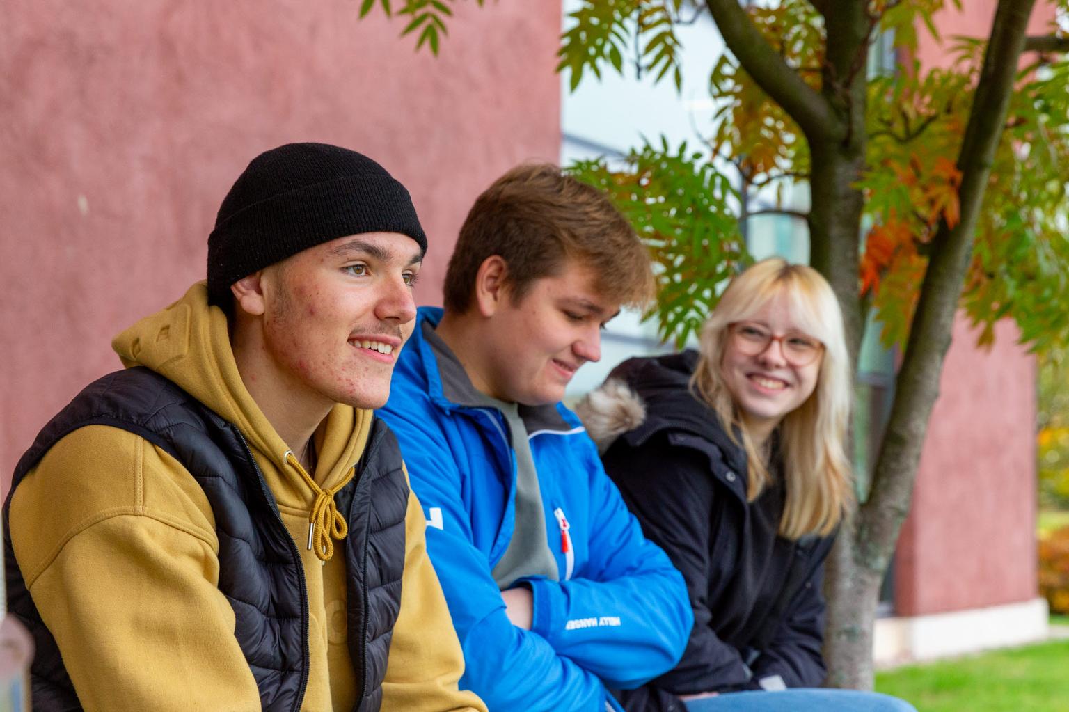 Three teenagers are sitting outdoors on a bench, dressed in warm jackets and hoodies, smiling and engaged in conversation. The background features a tree with autumn leaves and a red building, creating a cozy and relaxed atmosphere.