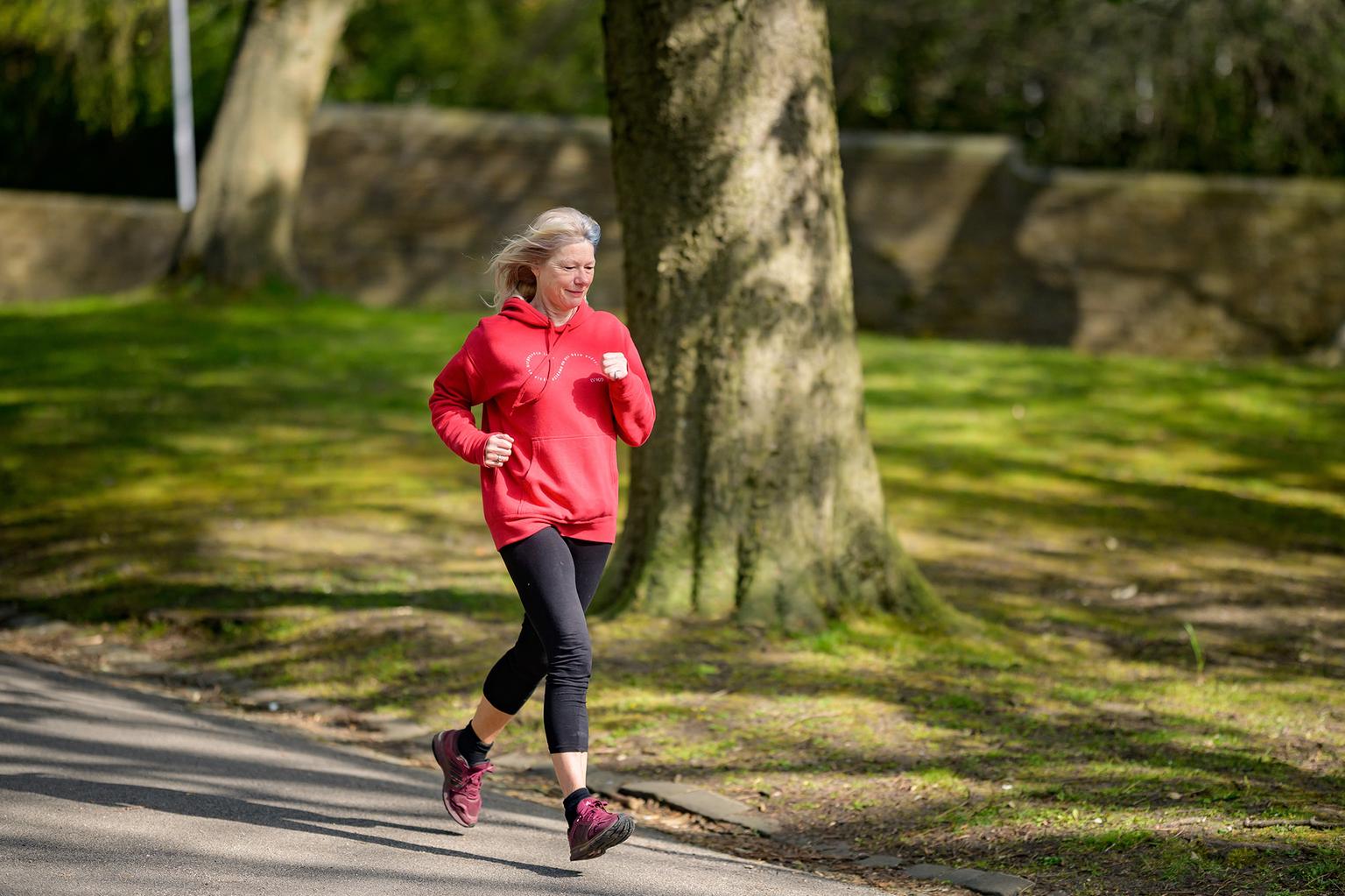 An older woman with silver hair is jogging in a park, wearing a red hoodie and black leggings. She looks focused and determined as she moves along a paved path surrounded by green grass and trees.