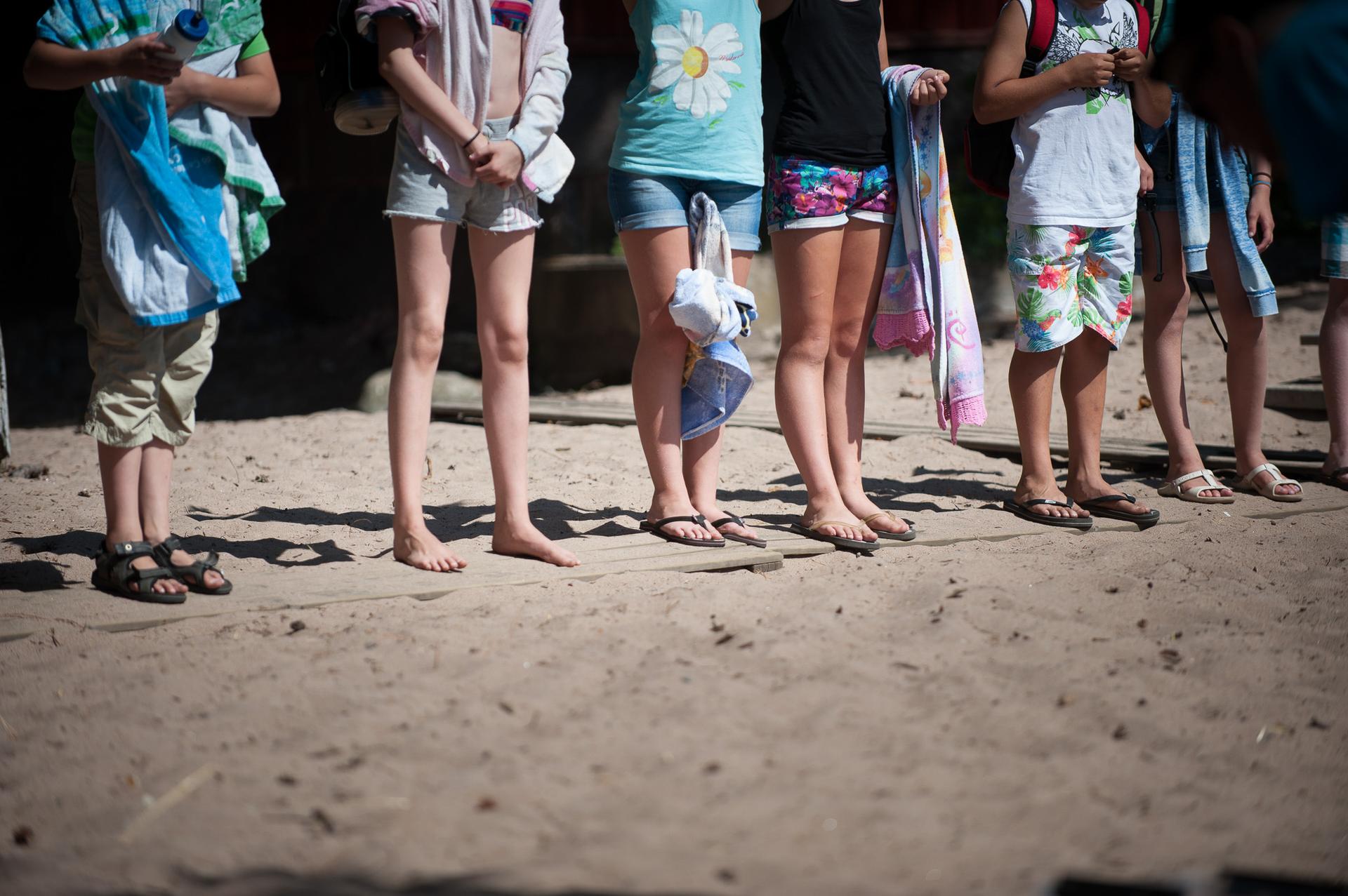 En grupp barn står på en sandstrand, vissa barfota och andra i sandaler. De håller handdukar och en vattenflaska, och ser ut att vara redo för bad eller en aktivitet vid vattnet.