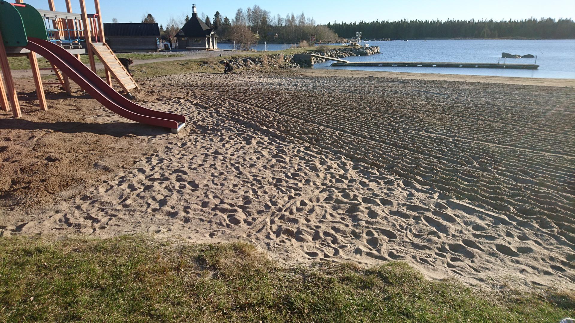 En lekplats vid en sandstrand med en rutschkana i förgrunden. Stranden är täckt av fotavtryck och har en brygga som sträcker sig ut i vattnet. I bakgrunden syns en grillstuga och en skog.