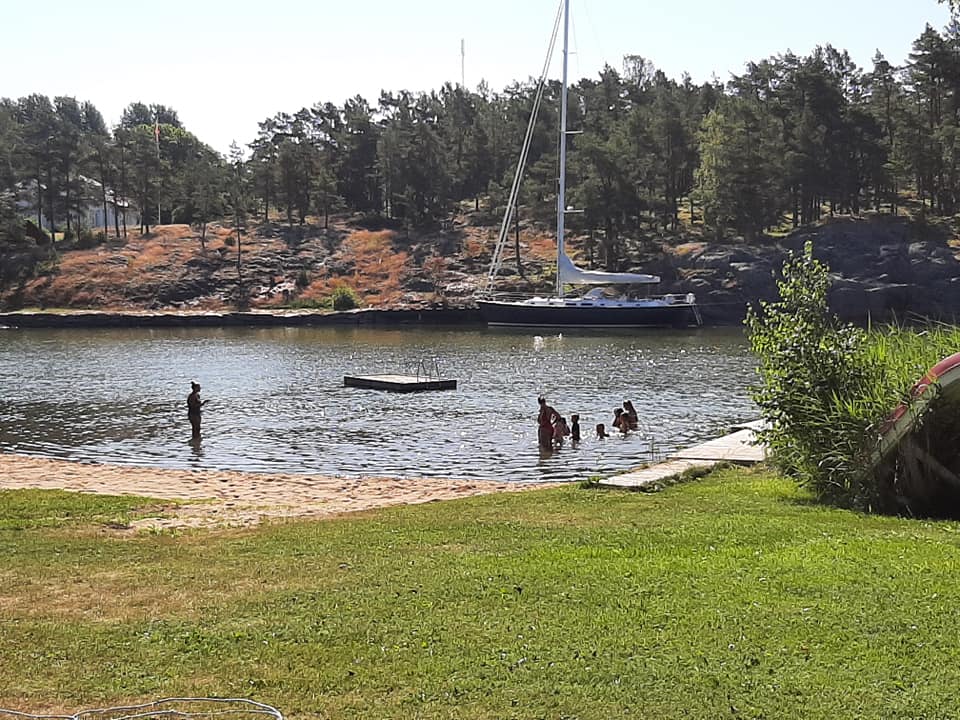En grupp människor badar vid en liten strand med en flytbrygga i vattnet. En person står längre ut i vattnet. I bakgrunden syns en segelbåt förtöjd vid en klippa och en skog med tallar.