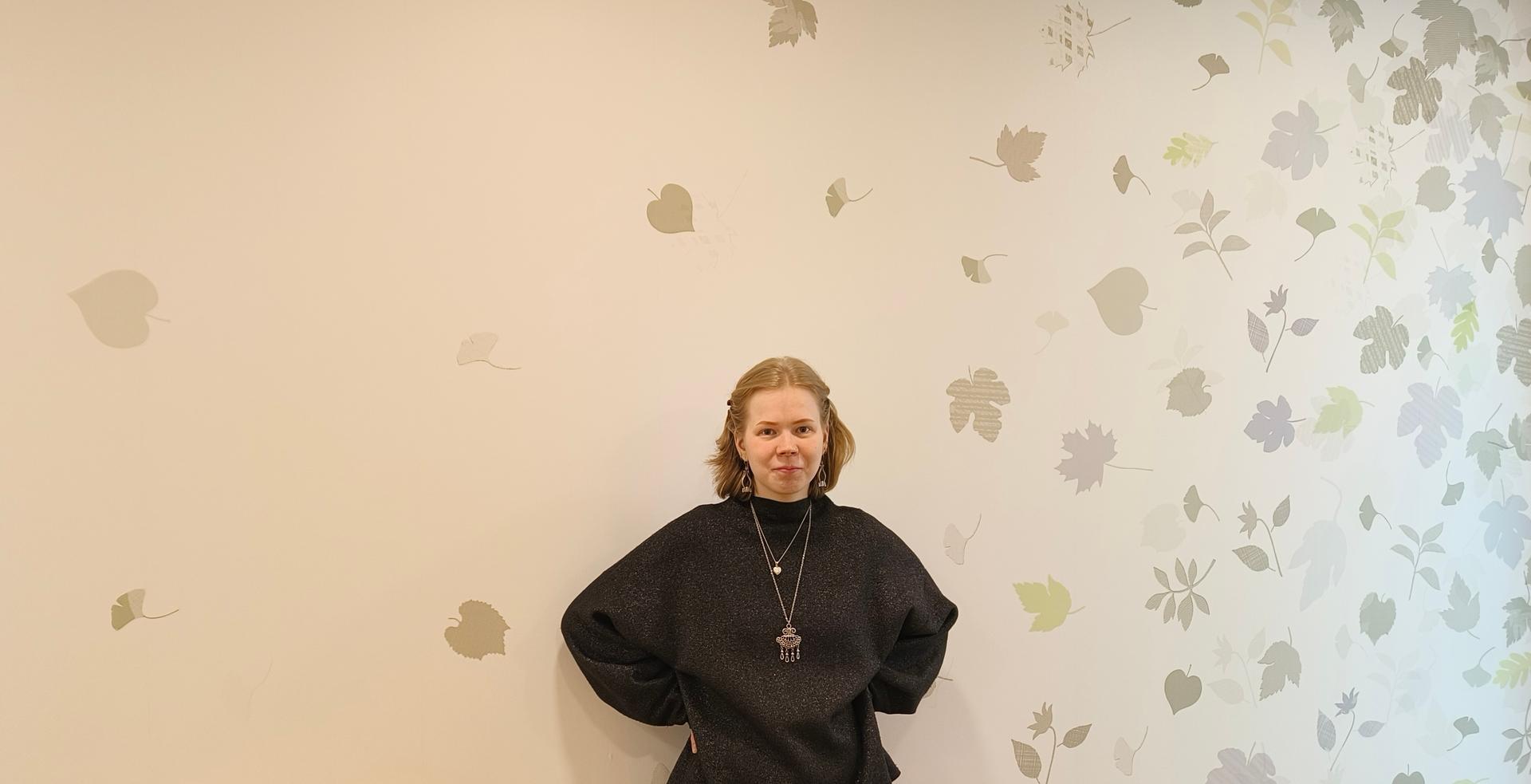 A person in a black top is standing in front of a light-colored wall decorated with leaf patterns. They are facing the camera with their hands behind their back.