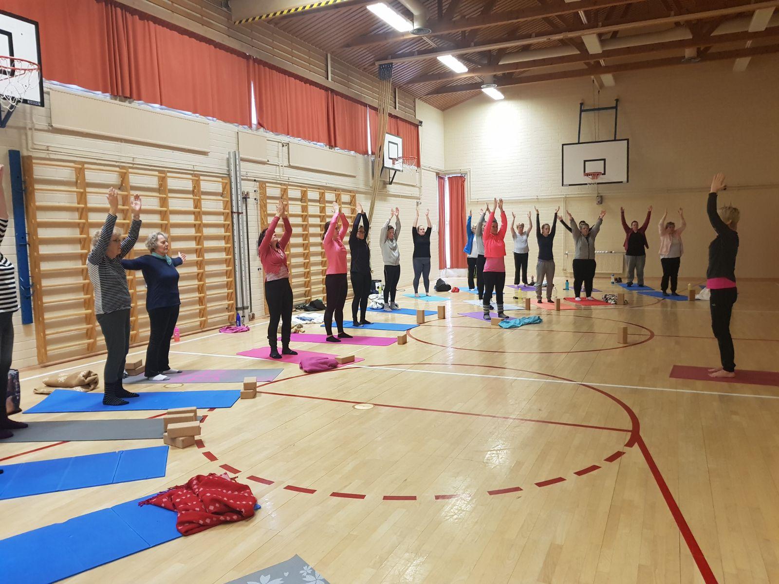 En grupp personer deltar i ett yogapass i en gymnastiksal. De står på varsin träningsmatta och sträcker armarna uppåt medan en instruktör leder övningen längst fram. Salen har ribbstolar på väggarna och basketkorgar i bakgrunden.