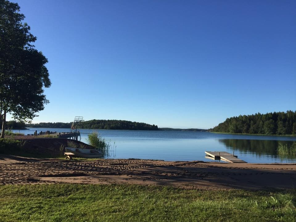 En stillsam sjö med bryggor och en sandstrand. En trampolin står vid en av bryggorna, och vattnet speglar den klarblå himlen. Träd och grönska omger området.