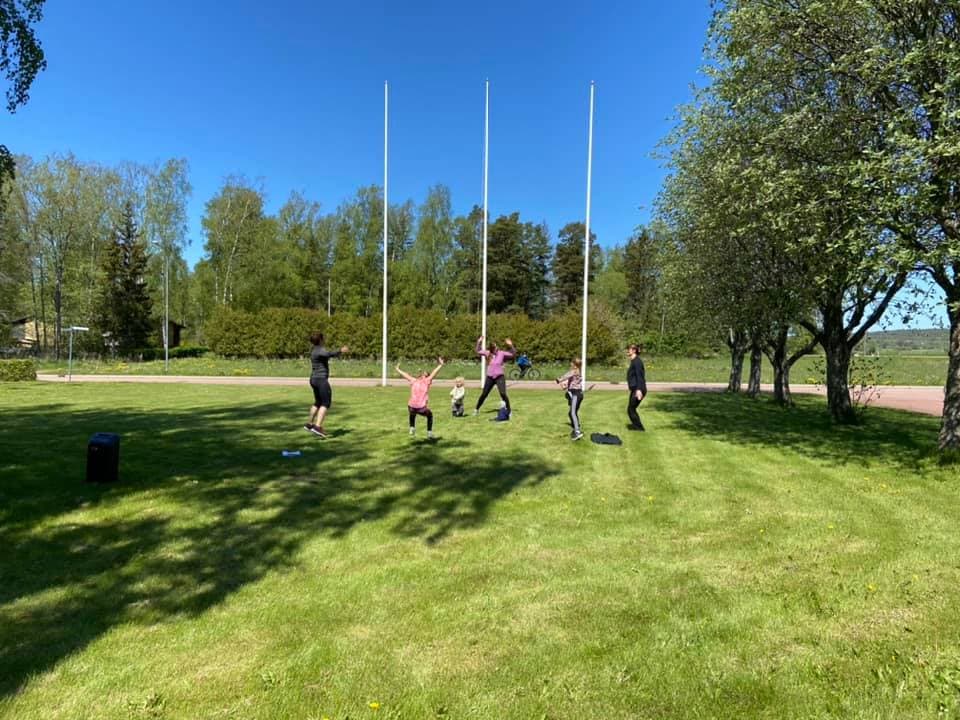 En grupp barn och vuxna deltar i en utomhusaktivitet på en gräsmatta med flaggstänger i bakgrunden. De rör sig med uppsträckta armar, och skuggorna från träden faller över gräset.