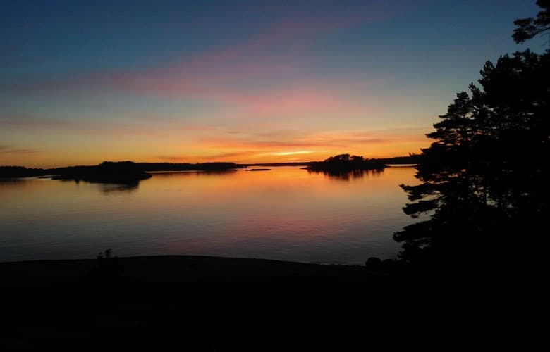 Solnedgång över en stilla skärgård, där himlen skiftar i varma nyanser av orange, rosa och blått. Silhuetten av träd och öar speglas i det lugna vattnet
