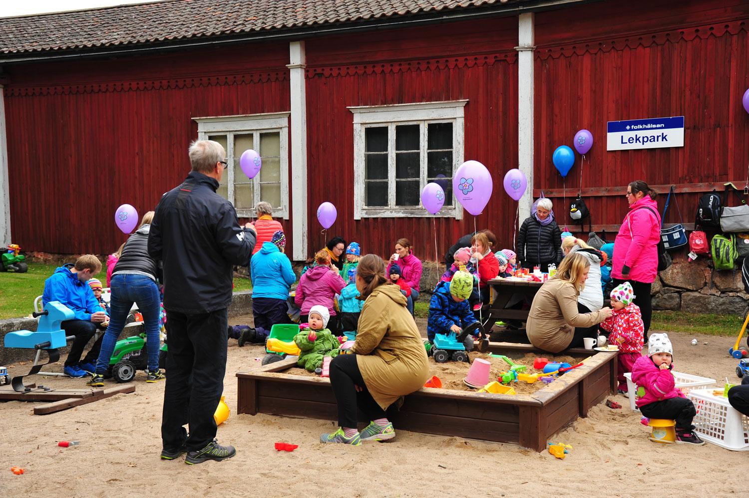 En lekpark utanför en gammal träbyggnad är fylld med barn och vuxna som deltar i olika aktiviteter. Barnen leker i sandlådan, kör leksaksfordon och håller ballonger, medan vuxna samtalar och hjälper till. En skylt på väggen visar att platsen är en Folkhälsan-lekpark.