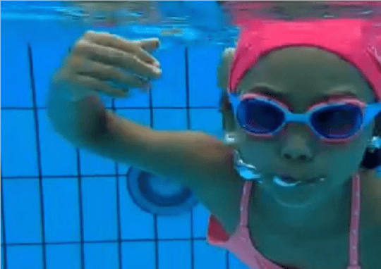 Ett barn med rosa badmössa och simglasögon simmar under vattnet i en pool. Bubblor syns kring munnen, och barnets arm är lyft i en simrörelse. Poolens kaklade botten och väggar syns i bakgrunden.