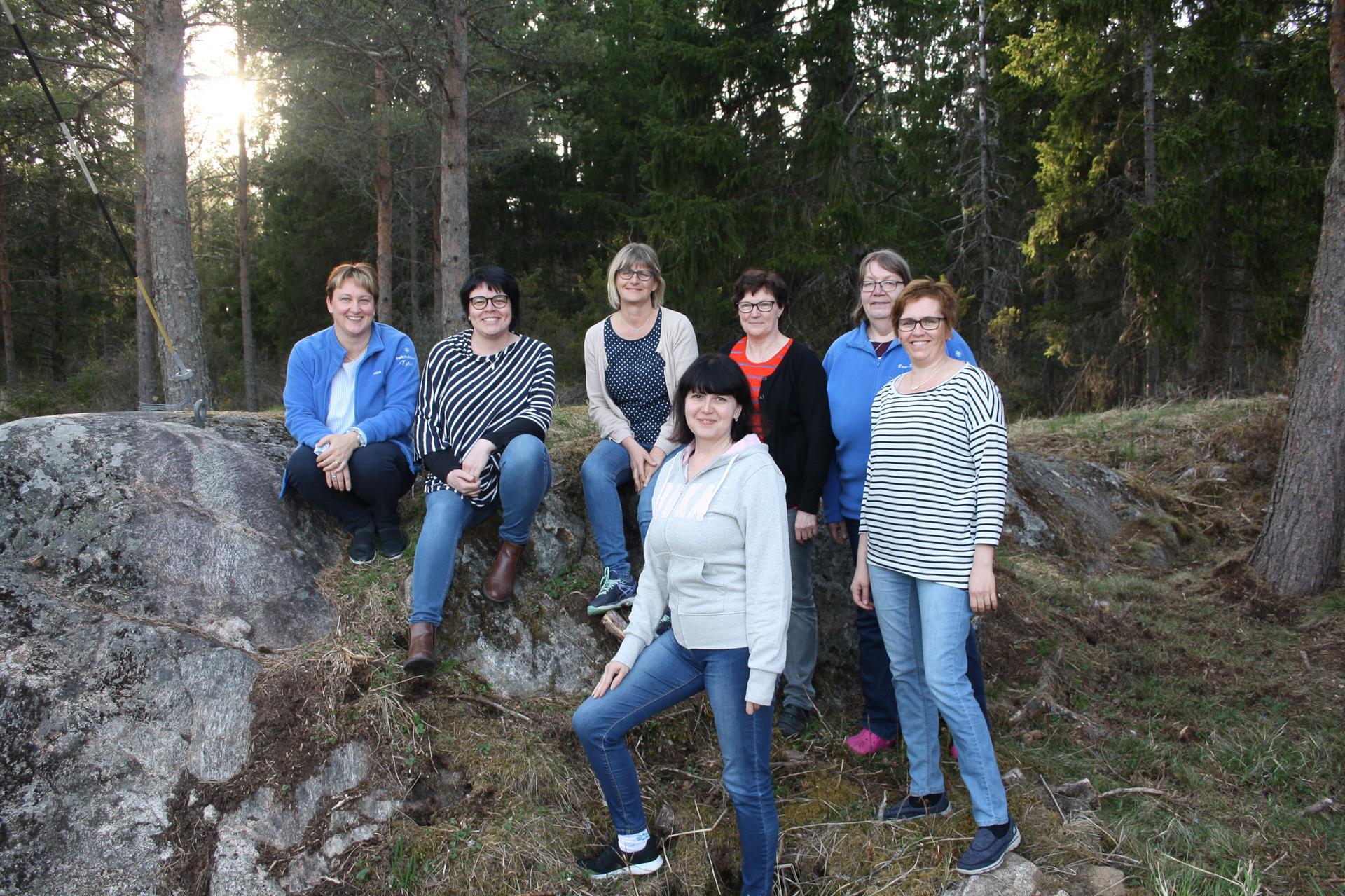 En grupp på sju personer poserar utomhus i en skogsmiljö. Några sitter på en sten medan andra står på marken. Bakgrunden består av träd och natur.