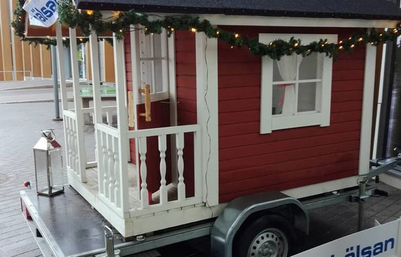 En röd lekstuga med vita detaljer står på ett släp dekorerat med ljusslingor och granris. Stugan har en liten veranda med vitt räcke och ett litet fönster med gardiner. I bakgrunden syns en stadsmiljö.