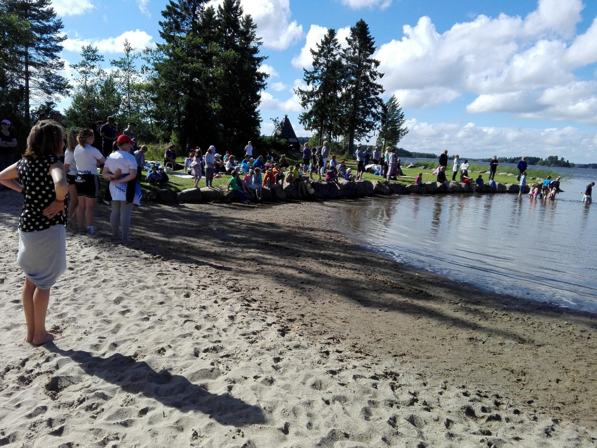 En stor grupp människor, både barn och vuxna, samlas vid en strand och på en gräsyta intill. Några står och tittar på medan andra sitter på stenar eller leker i vattnet. I bakgrunden syns träd och en liten byggnad.