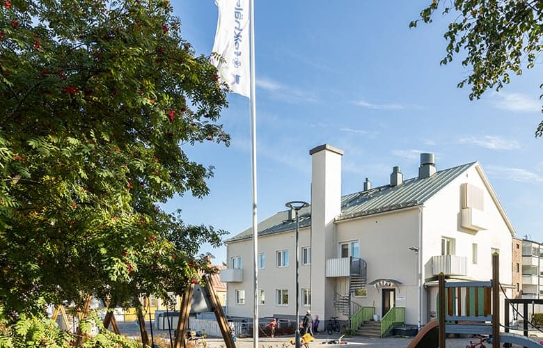 En byggnad med en tydlig fasad och detaljer syns i bilden. Framför huset finns en lekplats och en flaggstång med en hissad flagga. Himlen är klar och det är en solig dag.