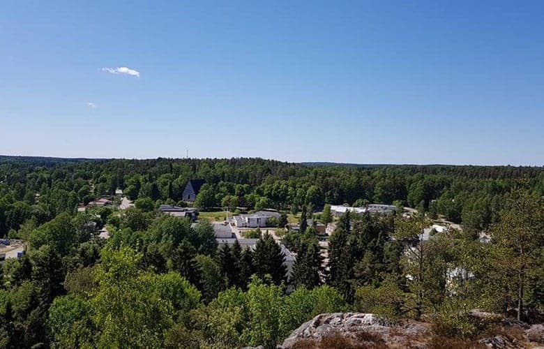 Vy över en grönskande småstad omgiven av skog. Bilden är tagen från en högre plats och visar bostadshus, en kyrka med en mörk takspets, samt vägar som slingrar sig genom det frodiga landskapet. Himlen är klarblå med enstaka moln, och naturen dominerar synfältet.