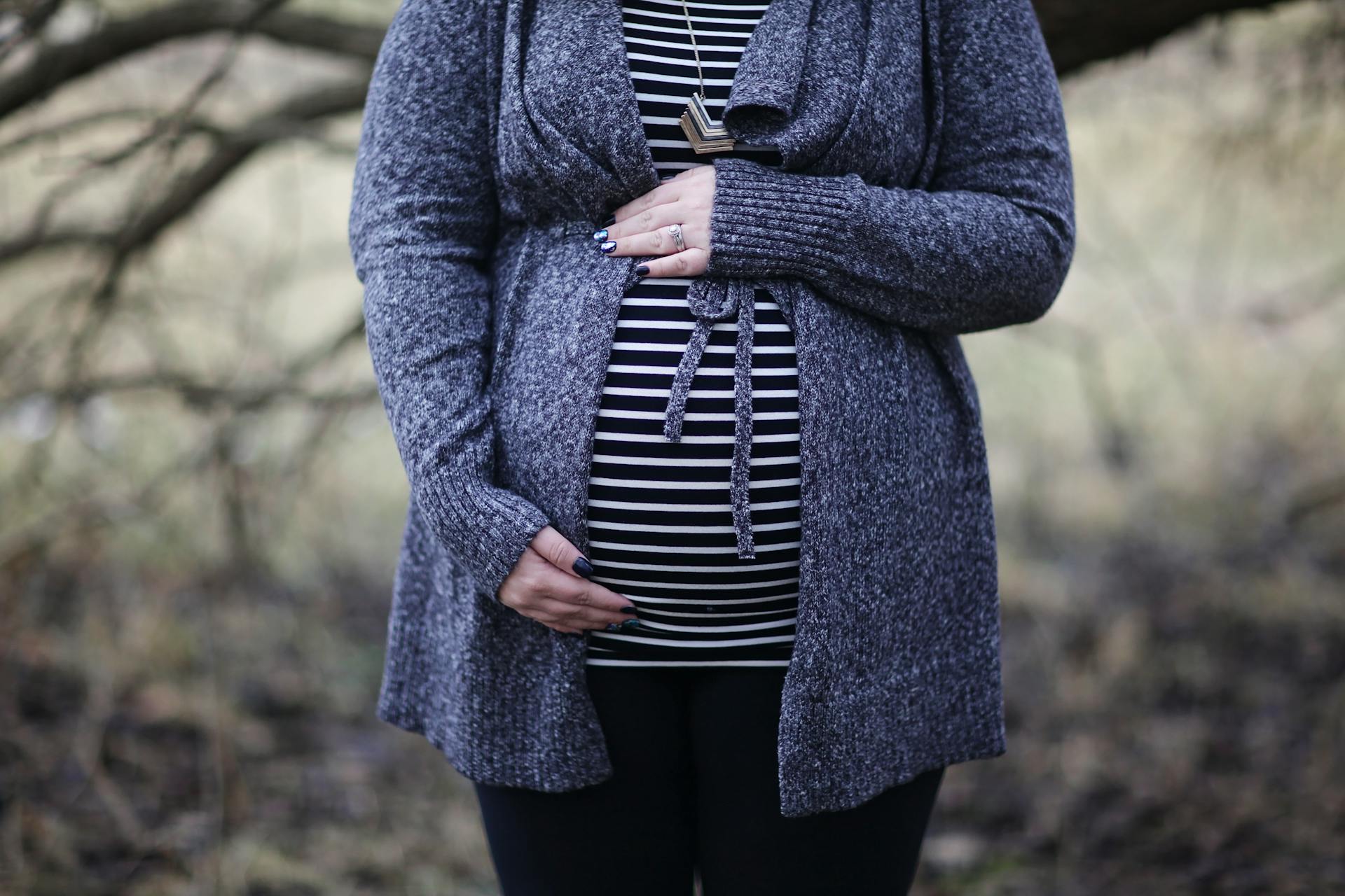 A pregnant woman stands outdoors, gently cradling her baby bump with both hands. She is wearing a striped top and a gray knitted cardigan, with a blurred natural background behind her.