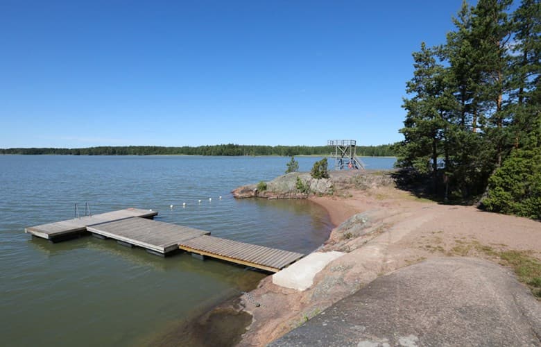 En naturskön badplats vid en sjö med en träbrygga som sträcker sig ut i vattnet. Klippor och tallar ramar in stranden, och en hopptorn står vid vattenkanten under en klarblå himmel.