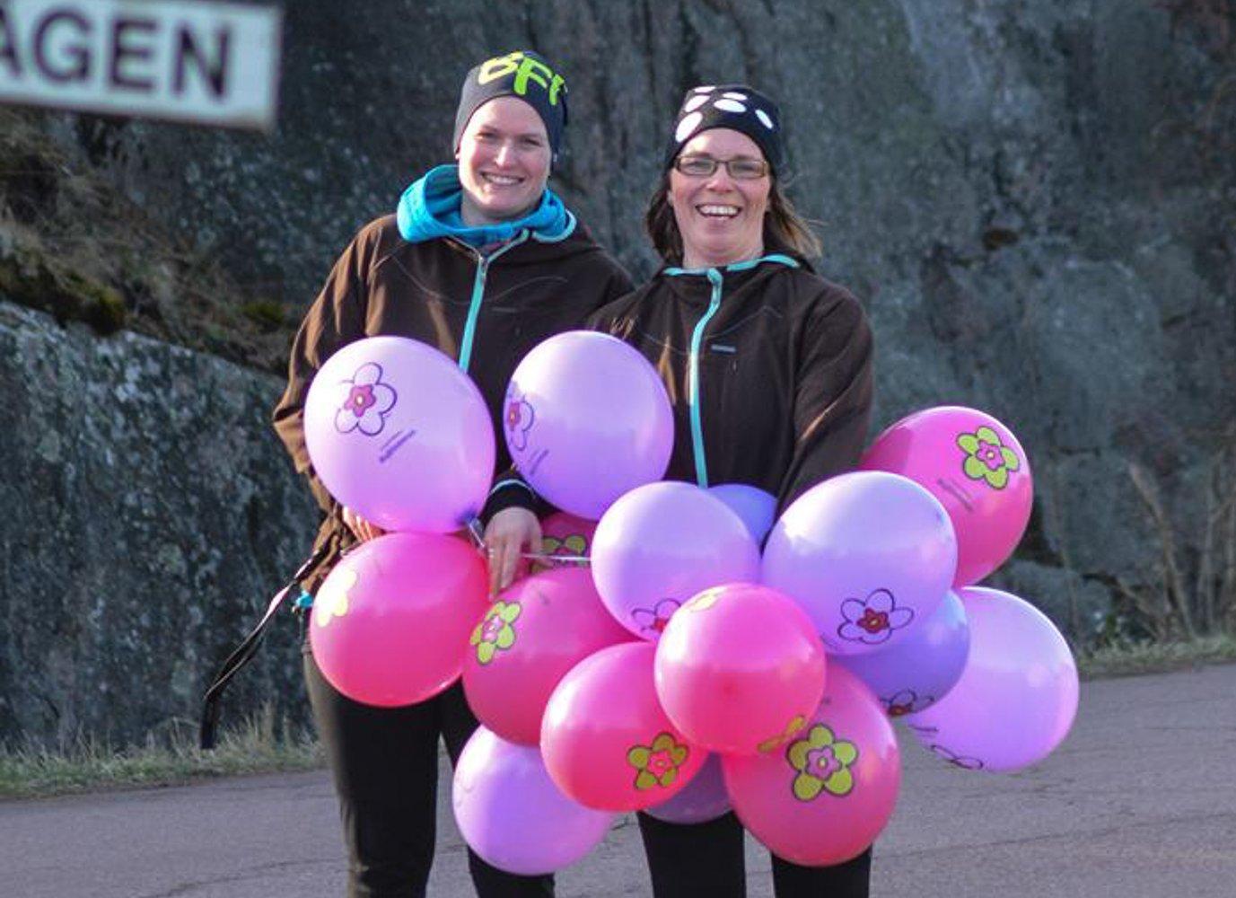 Två personer står utomhus vid en bergsvägg och håller en stor samling ballonger med blommotiv. De är klädda i varma kläder och mössor och ler mot kameran.