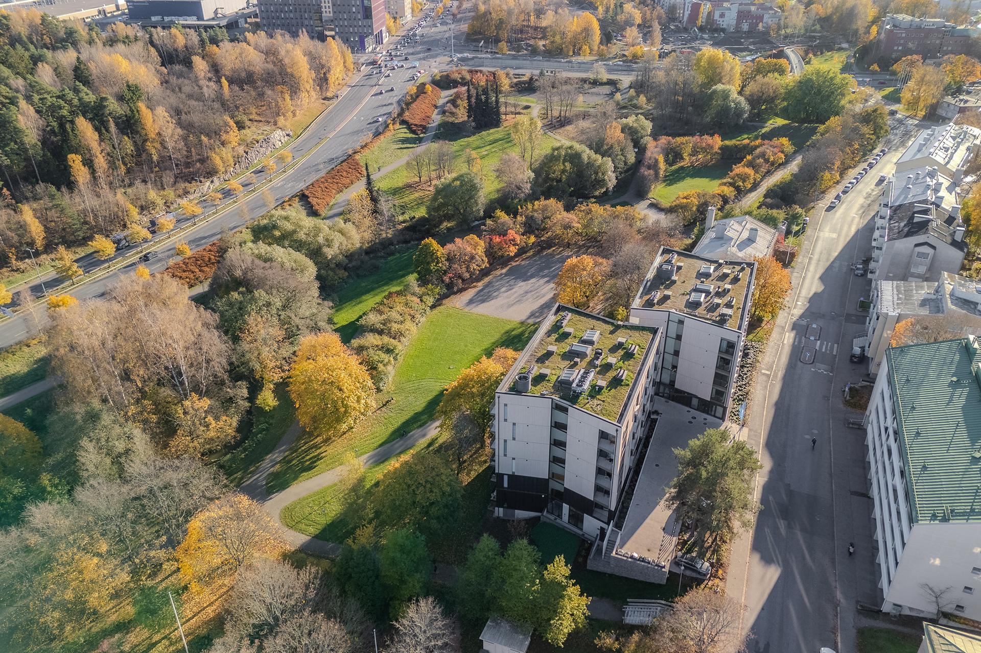 Flygbild över två moderna flervåningshus intill en grönskande park med promenadstigar och höstfärgade träd.