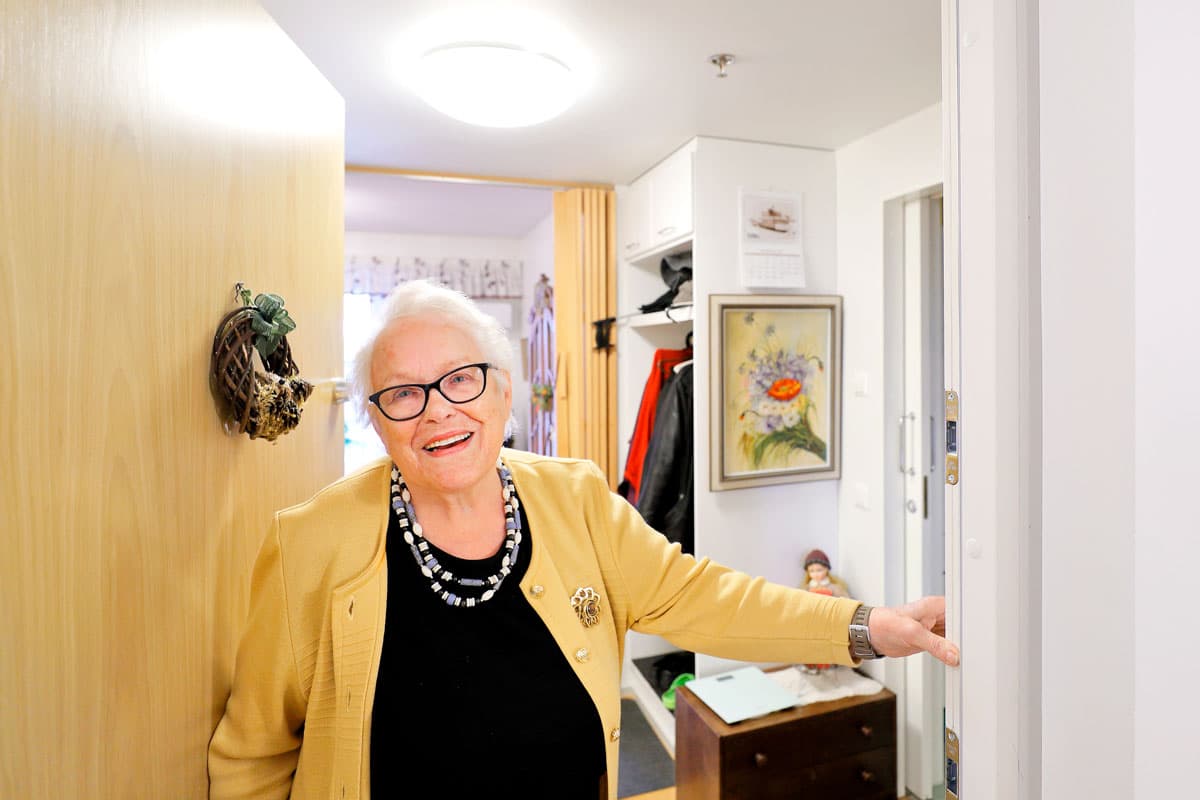 Vanhempi nainen, jolla on silmälasit ja keltainen jakku, seisoo kotinsa ovella hymyillen. Taustalla näkyy sisustusta ja seinällä oleva taulu.