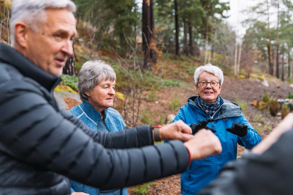 Tre äldre personer deltar i en aktivitet utomhus i en skogsmiljö. En kvinna i blå jacka leder eller instruerar gruppen medan de andra står samlade omkring henne bland tallar och höstfärgad vegetation.