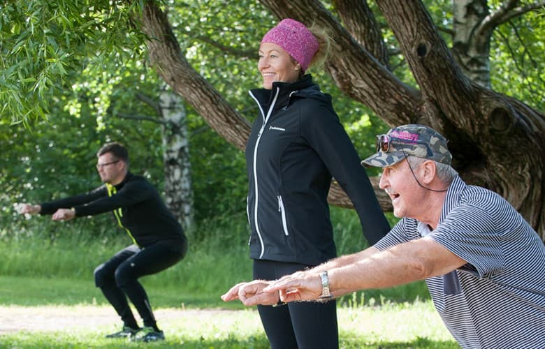 Tre personer motionerar i grupp utomhus. Personen närmast kameran gör knäböj.
