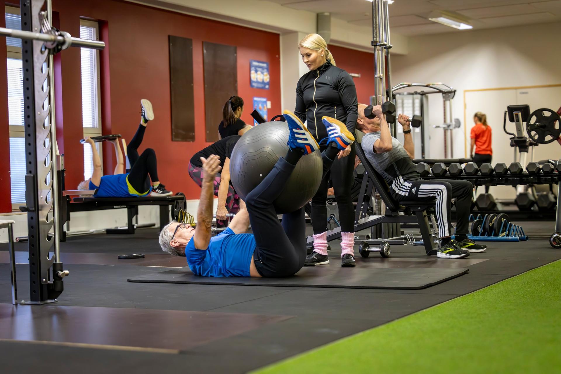 Ryhmä ikääntyneitä henkilöitä harjoittelee yhdessä valoisassa ja hyvin varustellussa kuntosalissa. Kaikki osallistuvat aktiivisesti ja ohjaaja tukee. Kuvassa välittyy yhteisöllisyys, liikkumisen ilo ja turvallinen, osallistava ilmapiiri.