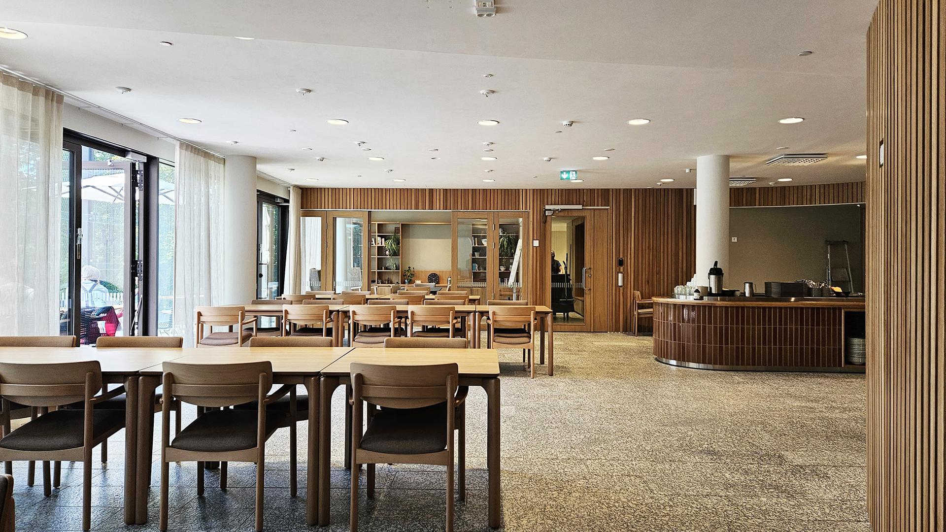 Ljus matsal med träbord och stolar, stora fönster, träpanel på väggarna och en serveringsdisk.