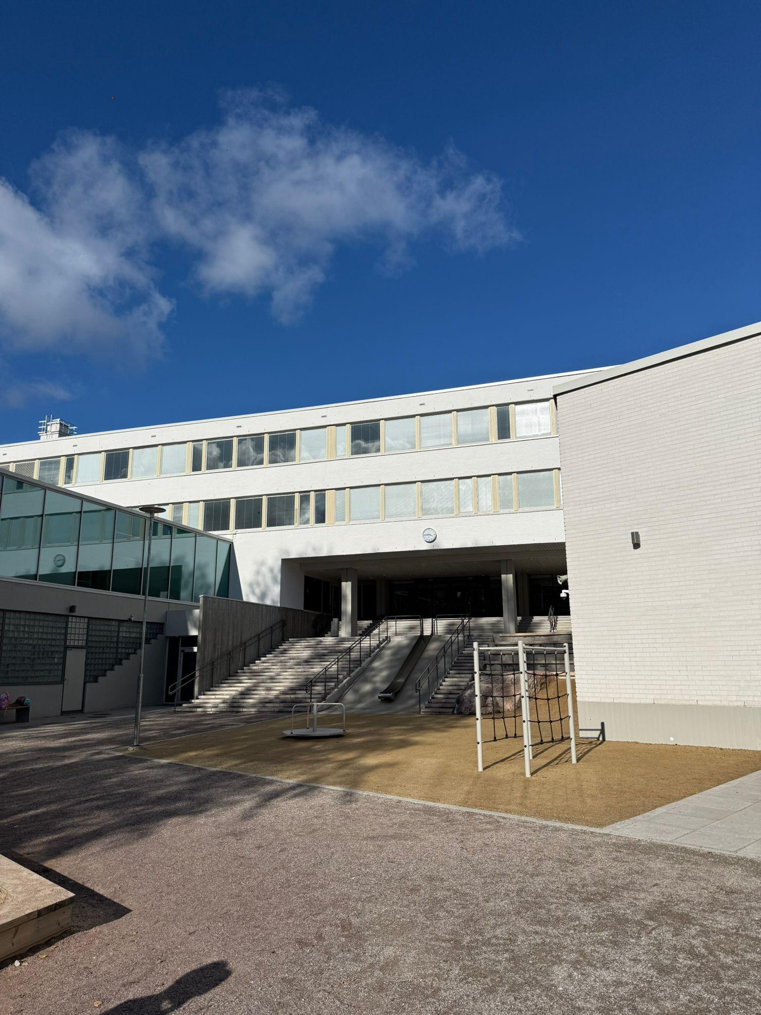 Koulupiha kiipeilytelineineen. Jykevät rappuset johtavat koulun sisäänkäyntiin.