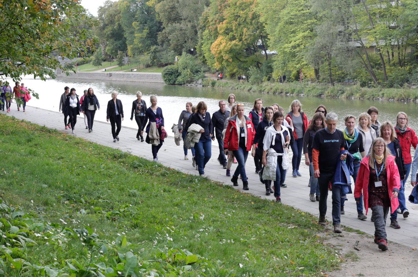 En stor grupp människor går längs en flodpromenad i en park under hösten. Personerna är klädda i jackor och tröjor, och många bär väskor eller skyltar runt halsen. Träden i bakgrunden har gröna och gula höstlöv.  