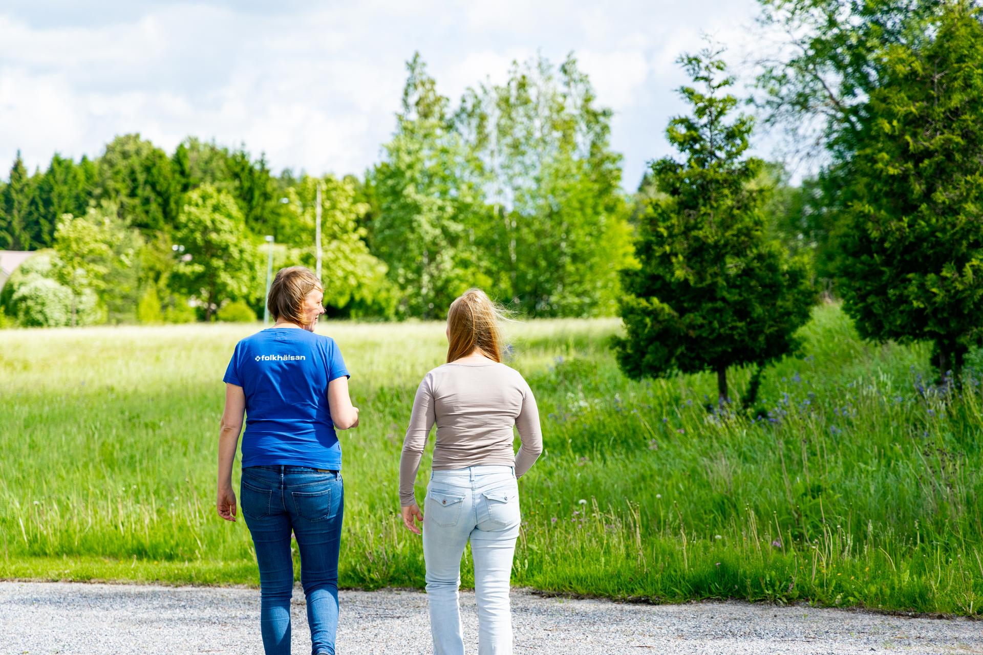 Två personer promenerar tillsammans på en grusväg i en grön naturmiljö en solig dag.