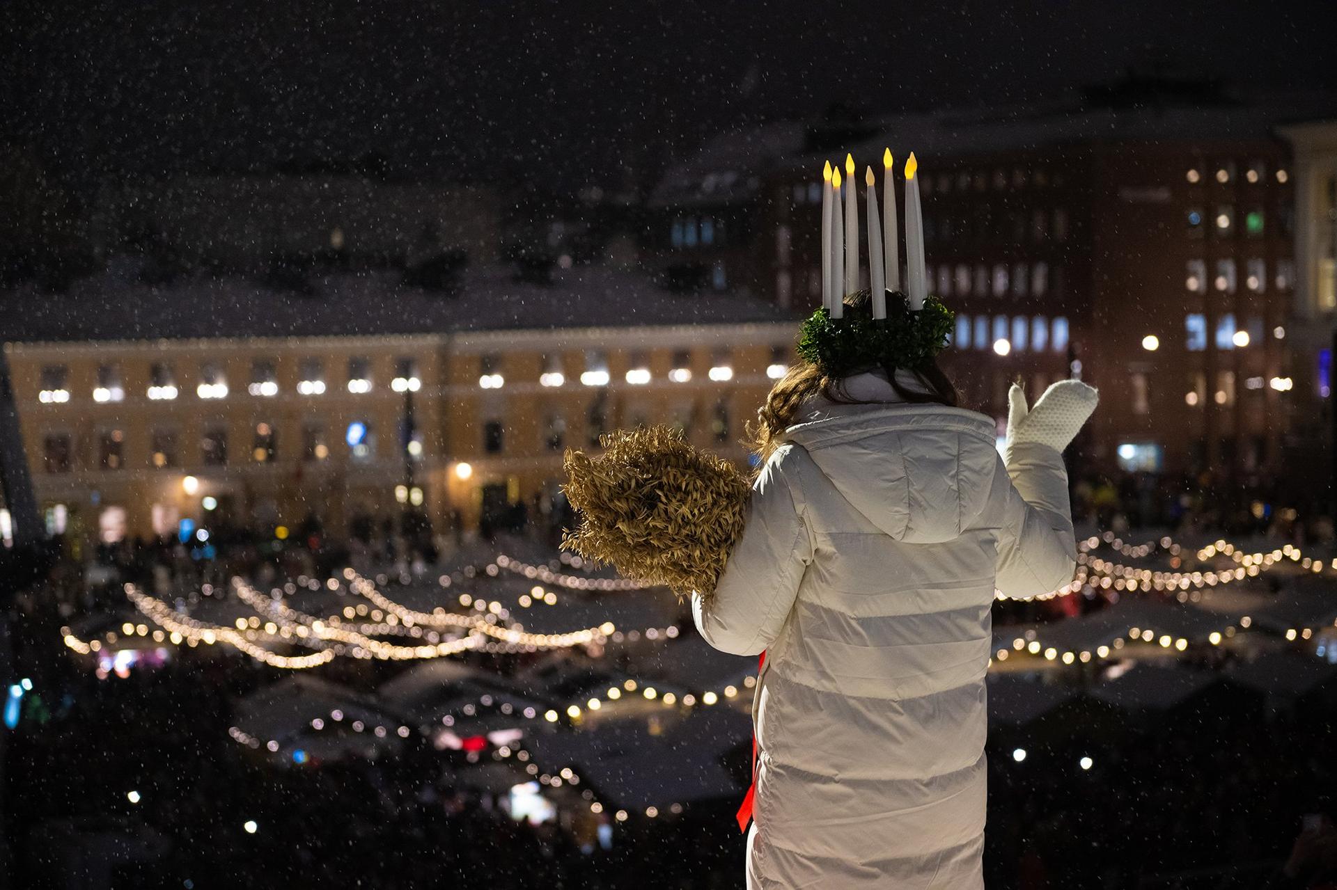 Finlands Lucia i vitt jacka och elektrisk ljuskrona står vänd med ryggen mot kameran och blickar ut över Senatstorget.