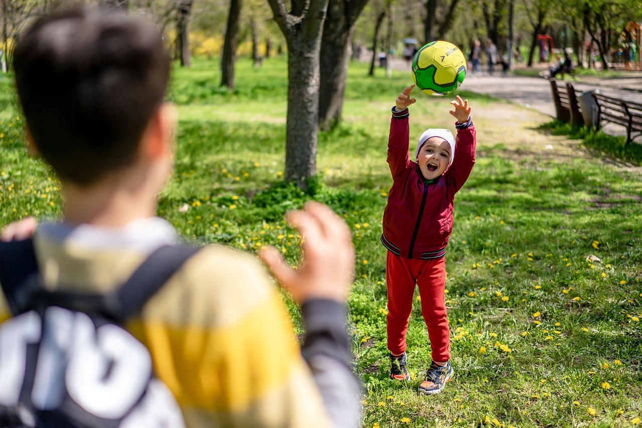 Ett barn kastar en boll och skrattar glatt i en solig park, medan ett annat barn står i förgrunden – en lekfull stund i trygg gemenskap.