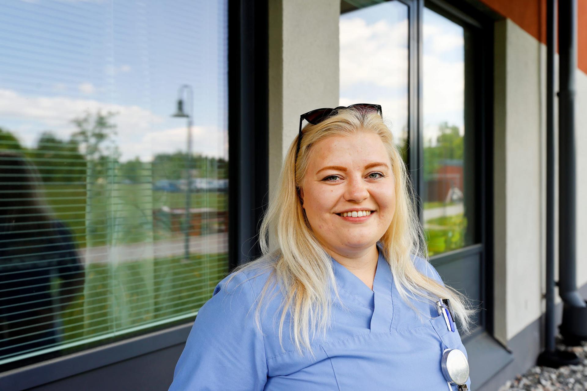 Hälsovårdare Josefine Fröberg står utomhus vid en byggnad med stora fönster och reflekterande glas. Hon bär ljusblå vårdkläder, har solglasögon på huvudet och en penna fäst i bröstfickan. Hon ler mot kameran, och i bakgrunden syns grönska och en lyktstolpe.