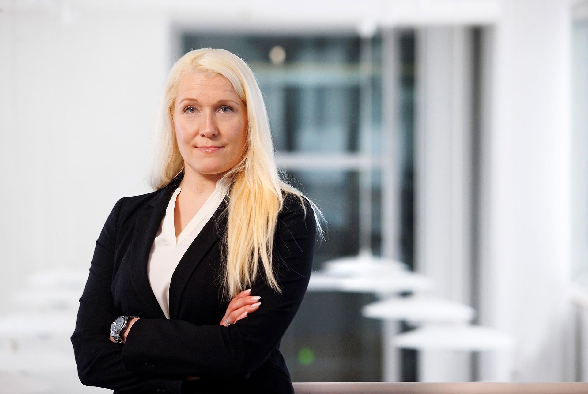 Portrait of a woman with long blonde hair standing with her arms crossed, wearing a black blazer and a white blouse. She is in a bright, modern indoor setting with a blurred background.
