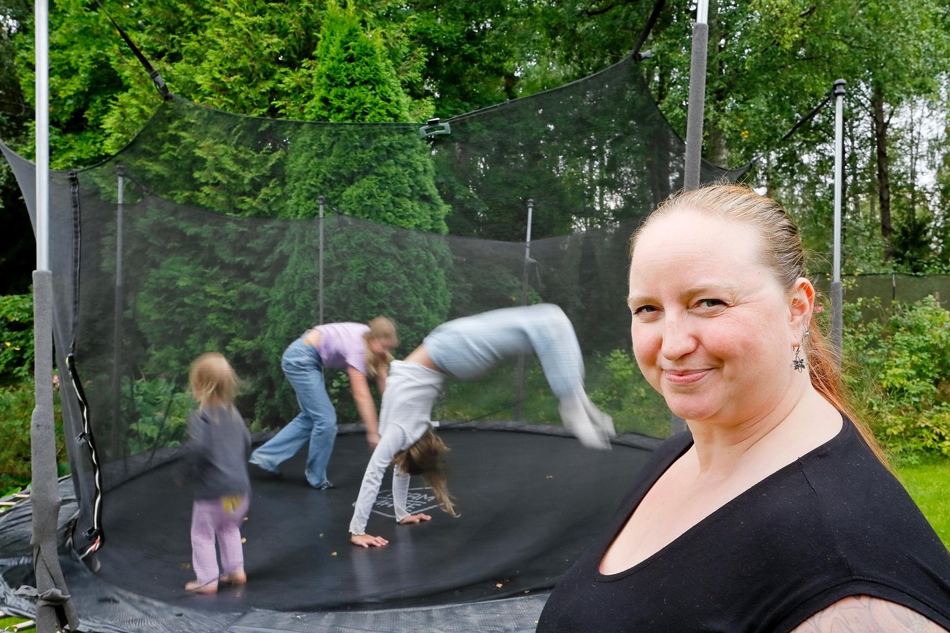 En mamma klädd i svarta kläder ser på sina tre barn som hoppar i trampolinen.