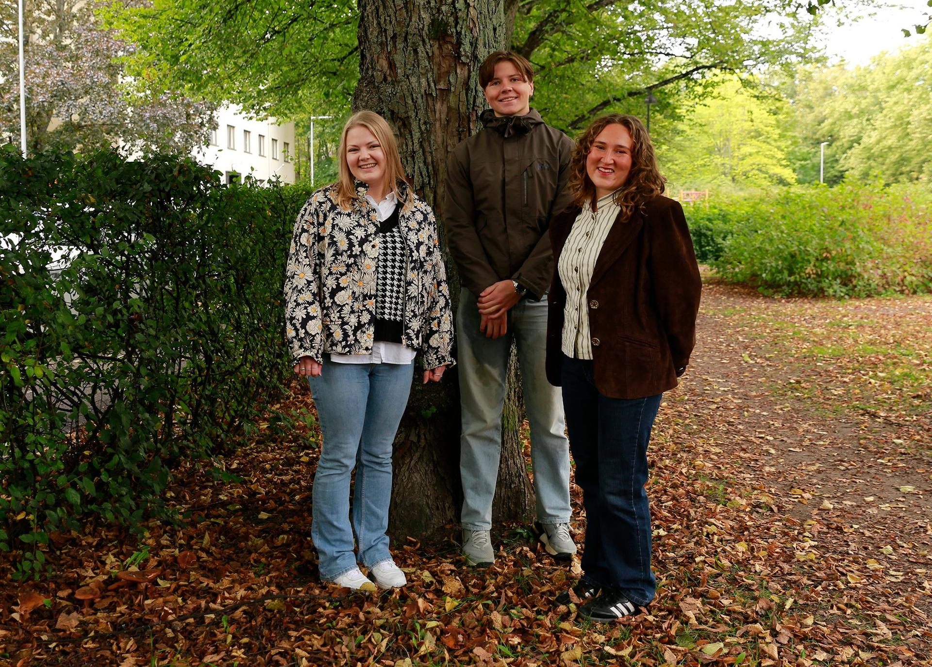 Tre unga vuxna står framför ett stort lummigt träd och poserar bredvid varandra. 