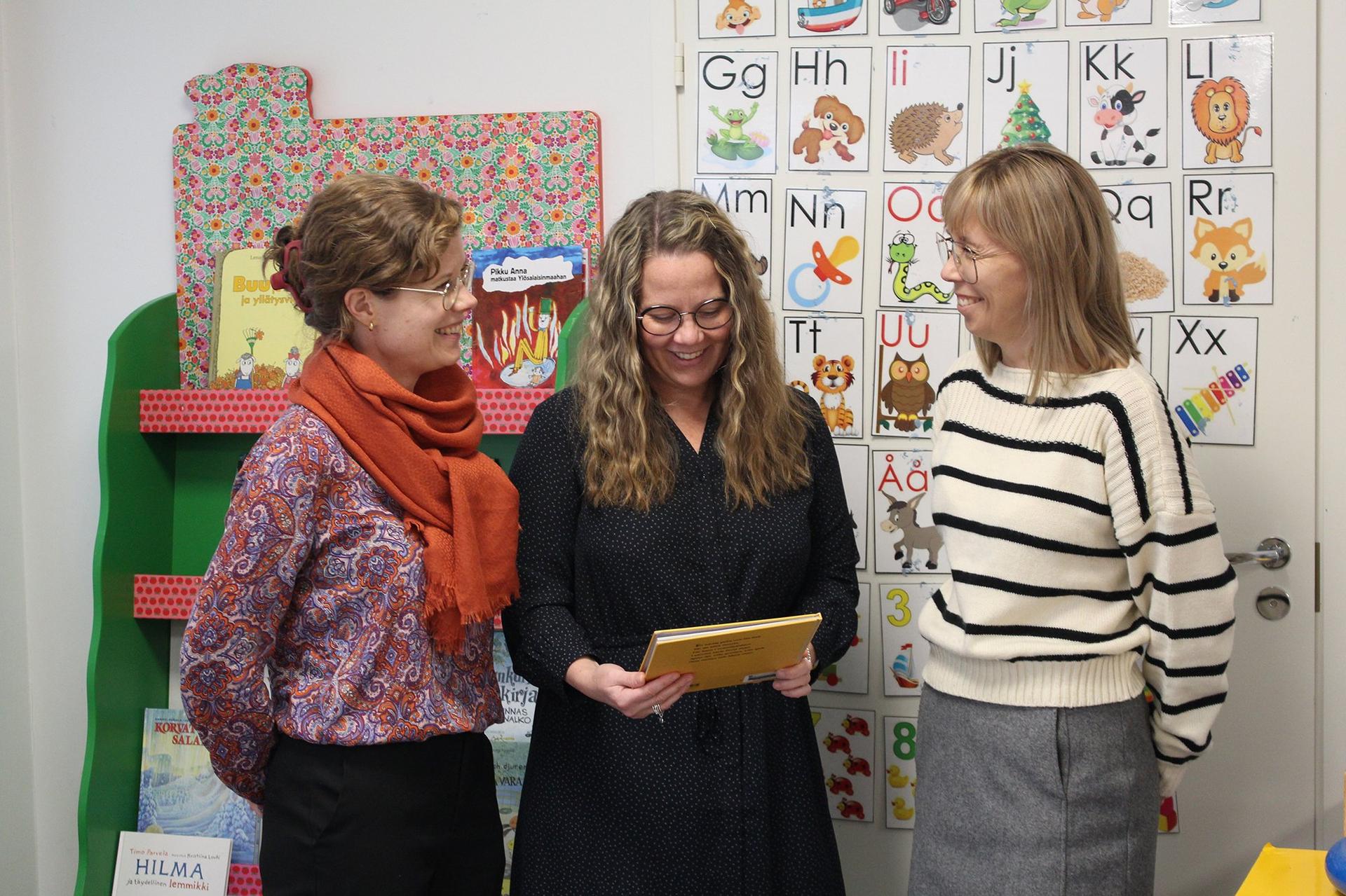 Tre kvinnor står brevid varandra och ser glatt på en barnbok. 