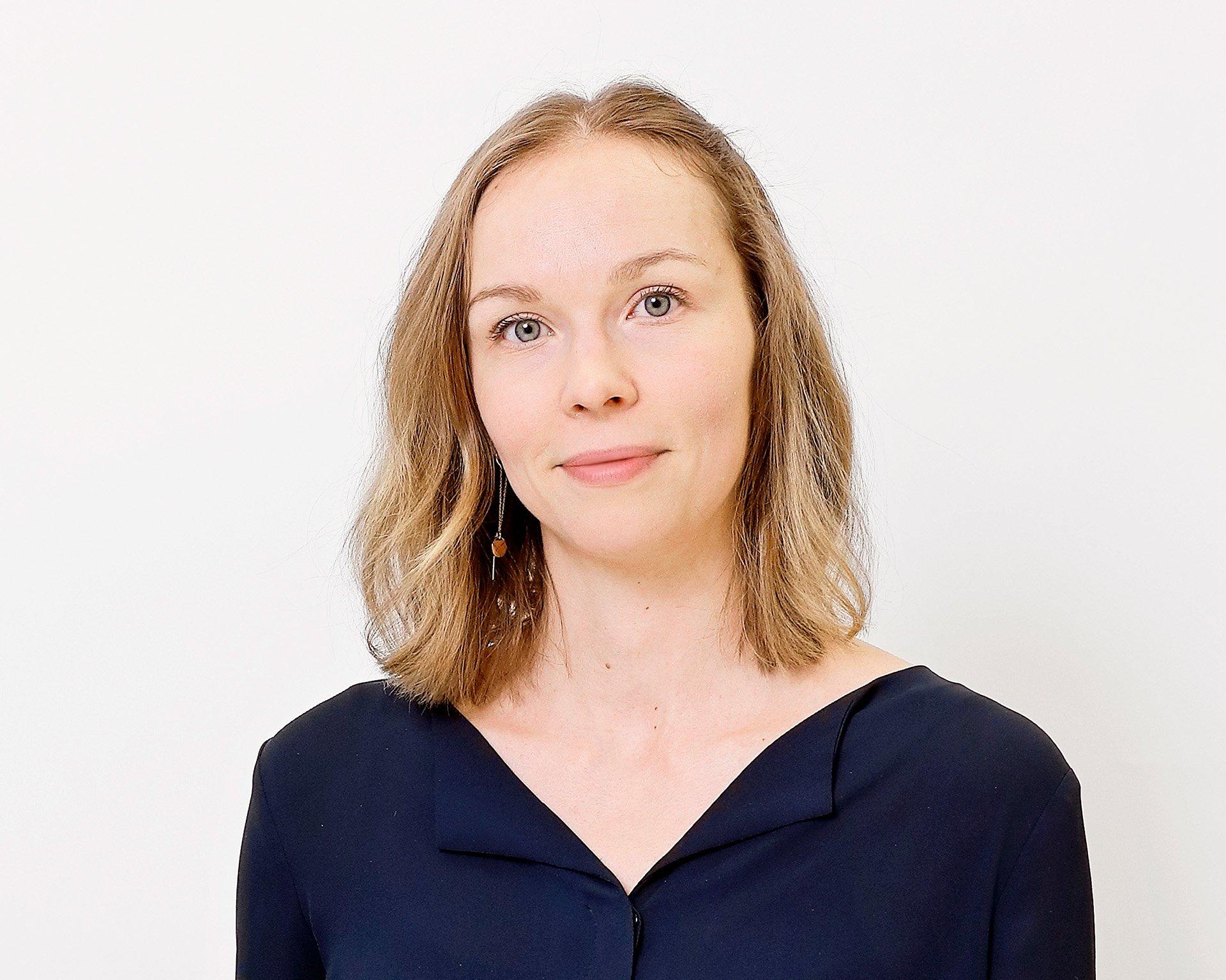 A woman with shoulder-length blonde hair wearing a dark blue blouse poses against a white background. She looks calmly into the camera with a neutral expression.
