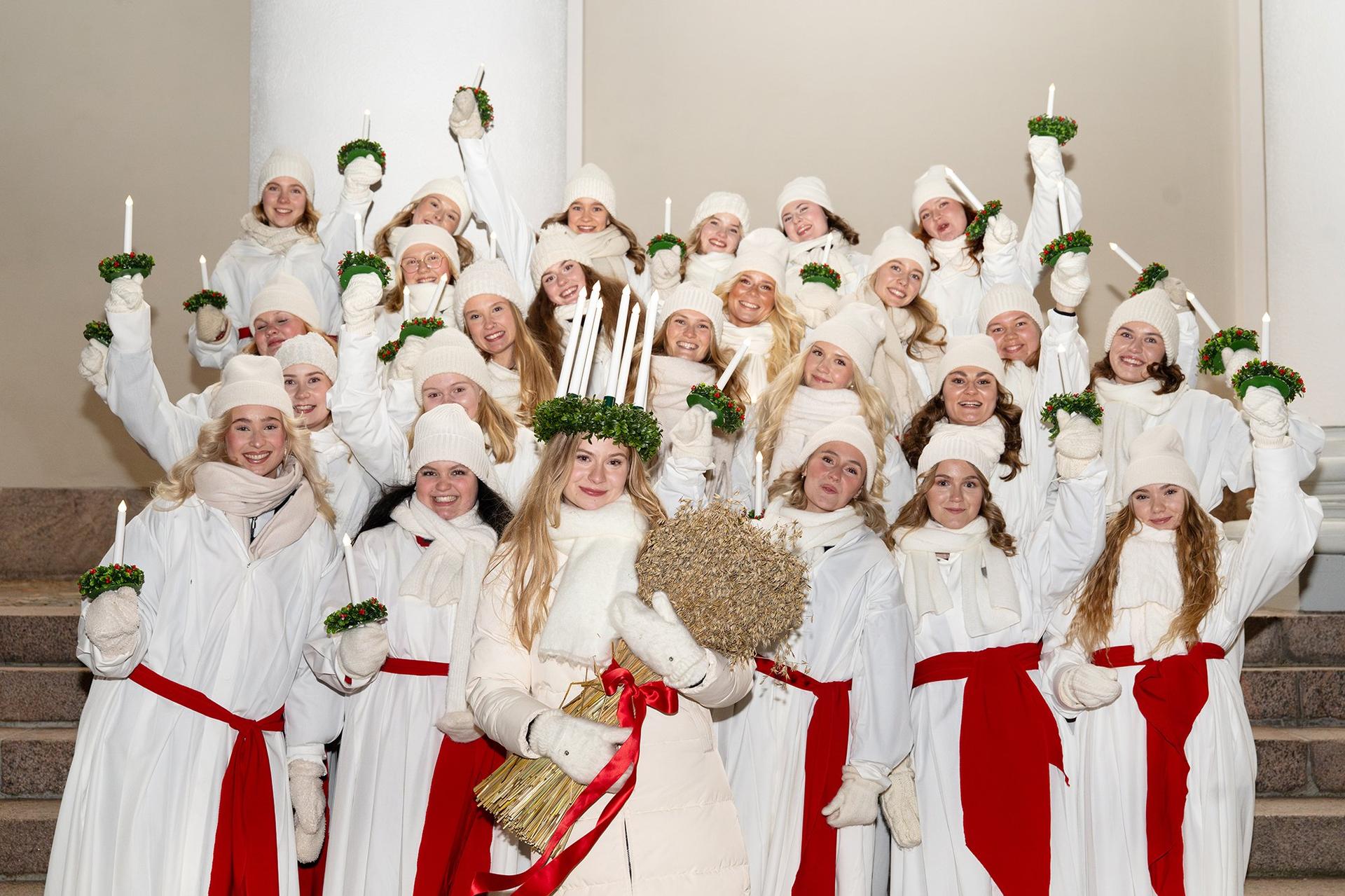 Finlands Lucia 2025 och Luciakören står tätt ihop framför en kyrkvägg, alla ser glada ut och håller upp sina ljus. 