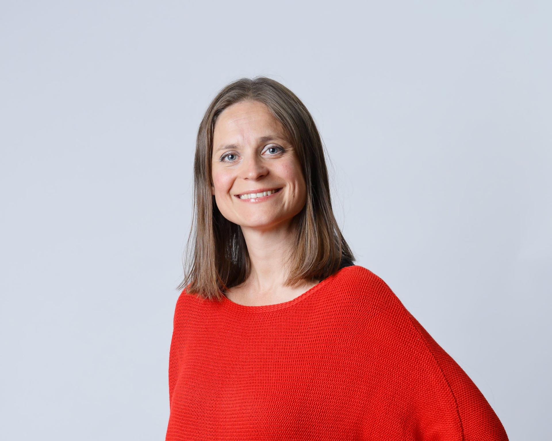 En person med axellångt brunt hår står vänd mot kameran och ler. Personen bär en röd tröja mot en ljus bakgrund.