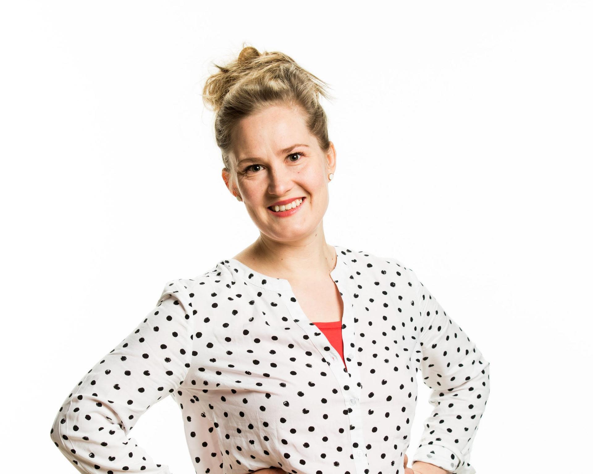 En person med uppsatt h&aring;r st&aring;r v&auml;nd mot kameran med h&auml;nderna placerade vid midjan. Personen b&auml;r en vit blus med svarta prickar och ett r&ouml;tt plagg syns under, mot en ljus bakgrund.