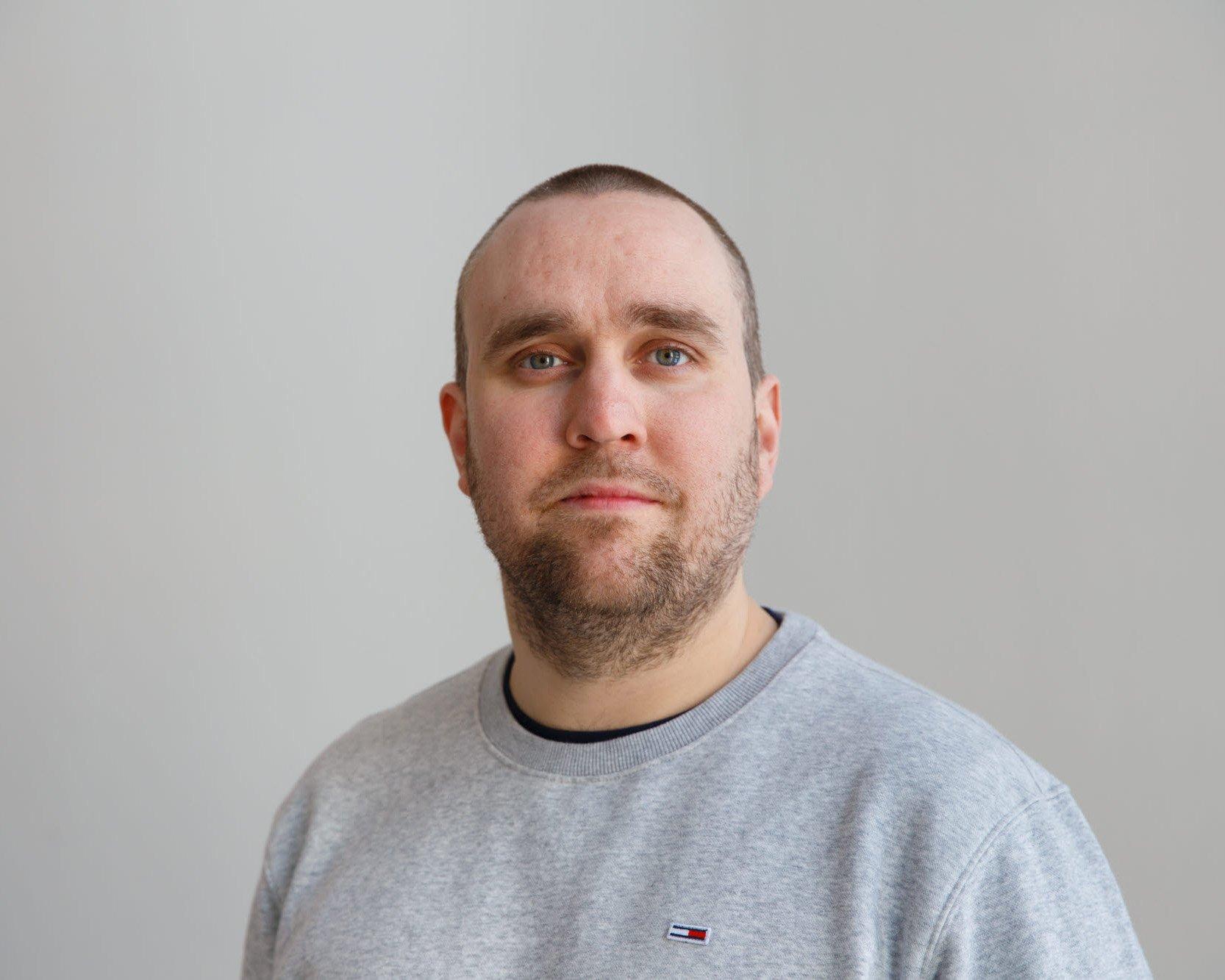 En person med kort hår och skägg står vänd mot kameran. Personen bär en ljusgrå tröja med en liten logotyp mot en ljus bakgrund.