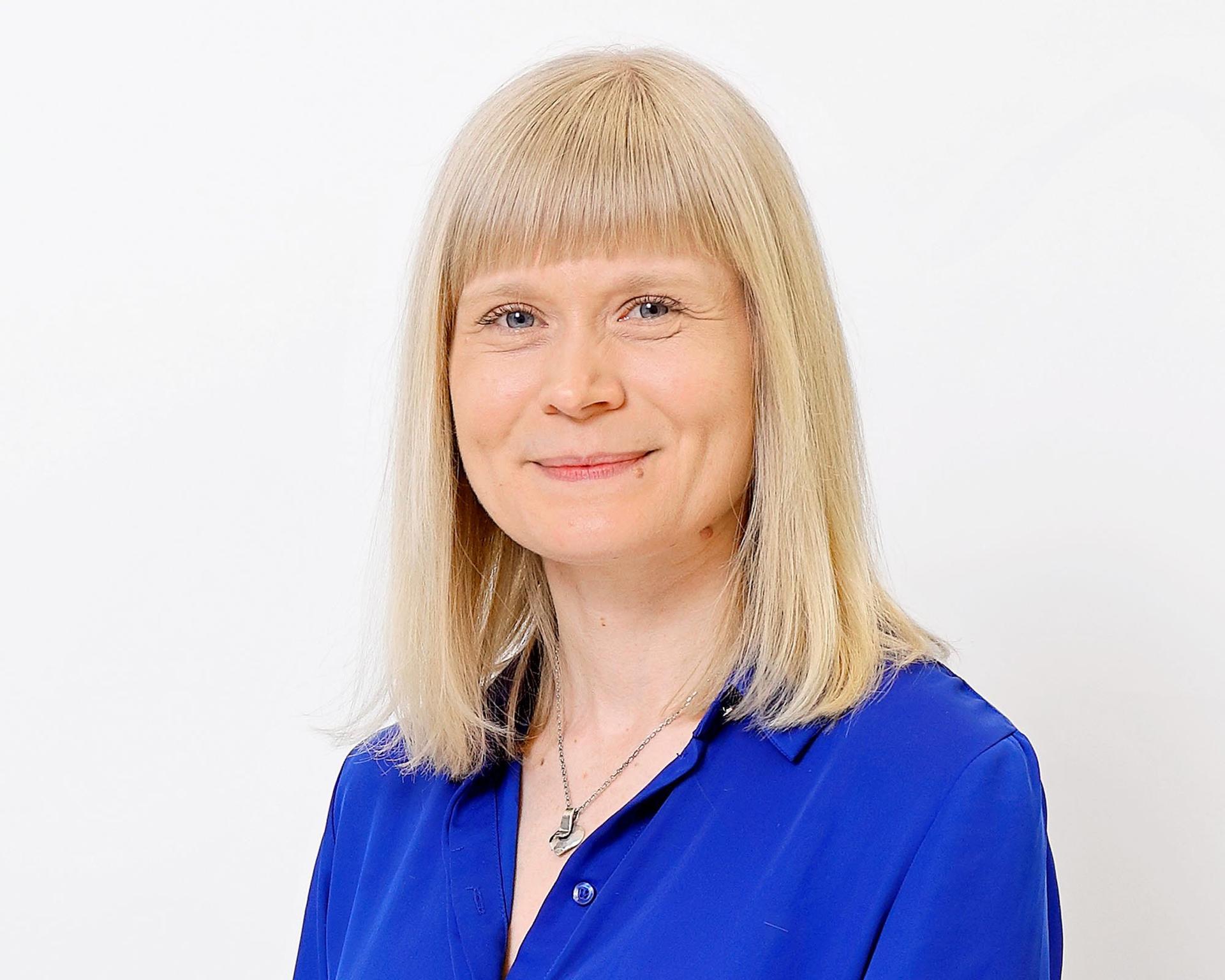 En person med axellångt ljust hår och rak lugg står vänd mot kameran. Personen bär en blå blus och ett halsband mot en ljus bakgrund.
