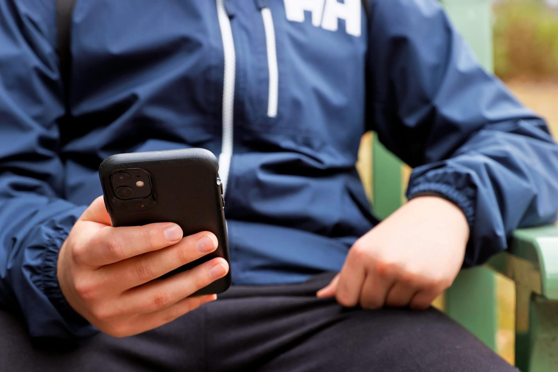 En ung person sitter ner och håller en mobiltelefon i ena handen. Personen bär en mörkblå jacka och sitter på en bänk med händerna synliga i bildens förgrund.