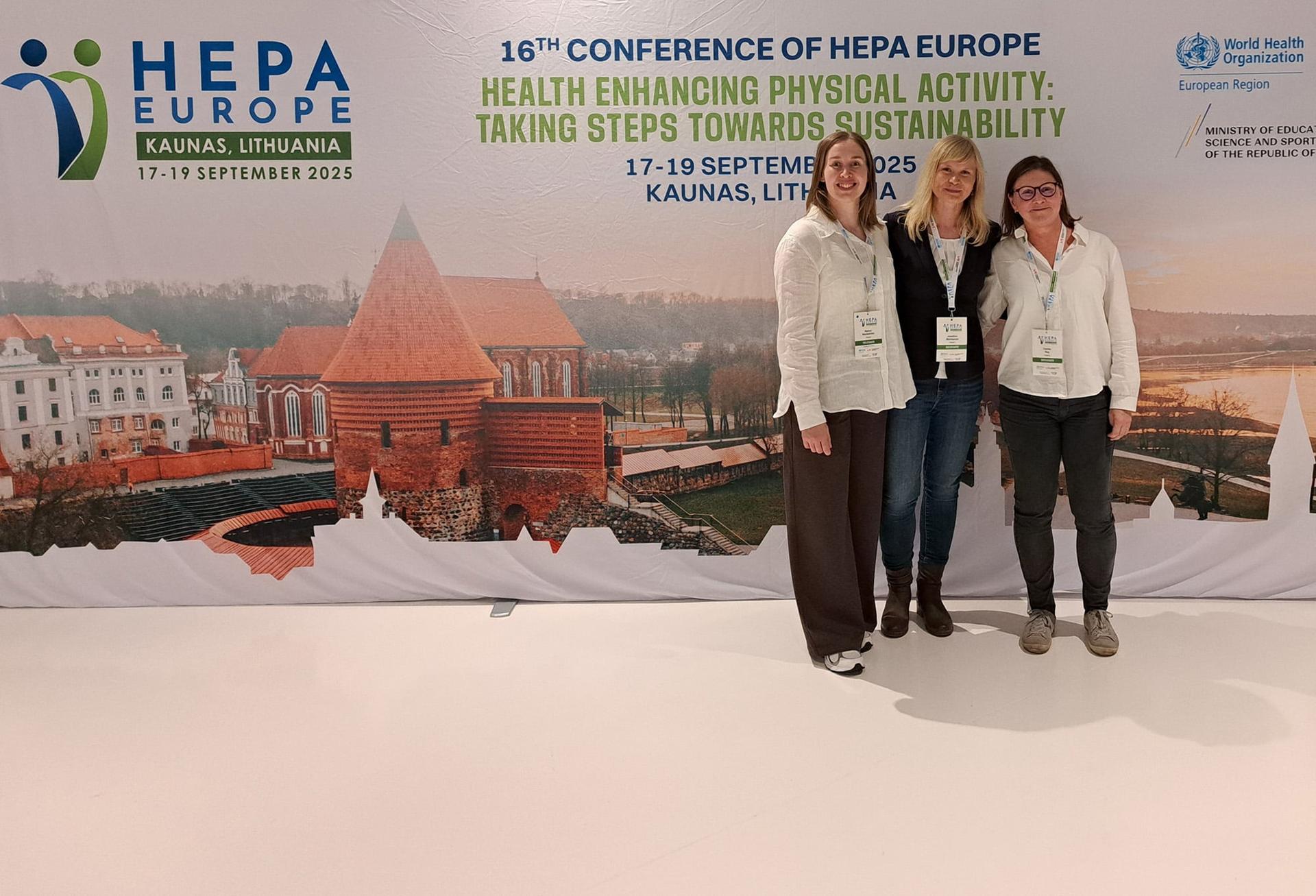 Tre personer står framför en stor konferensvägg för HEPA Europe i Kaunas, Litauen, med texten “Health Enhancing Physical Activity: Taking steps towards sustainability”. I bakgrunden syns en bild av Kaunas stadsmiljö och konferensens logotyper.