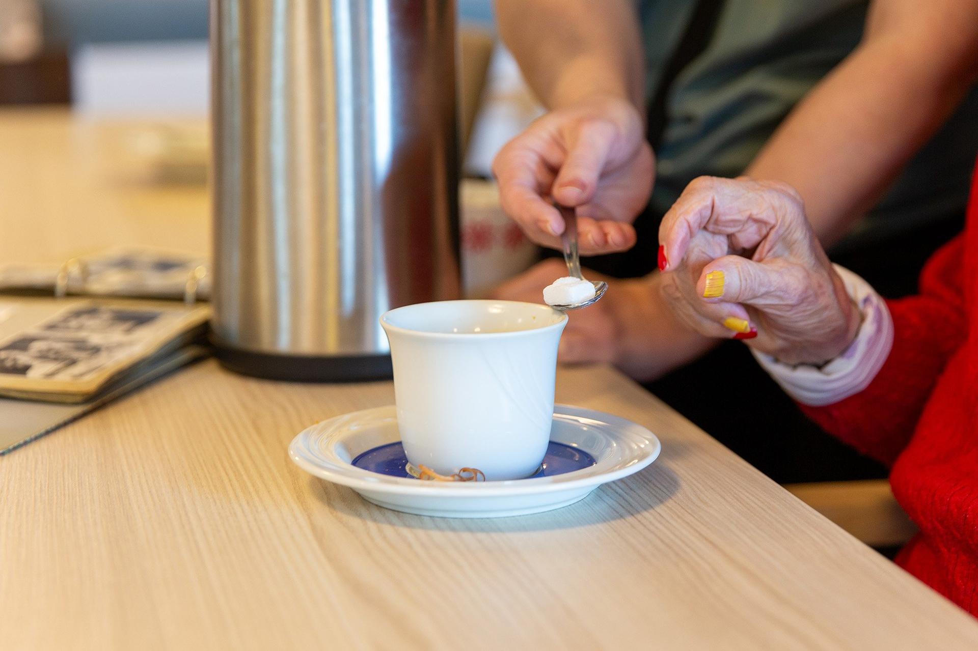 Äldre person får hjälp att lägga socker i en kopp kaffe vid ett bord.
