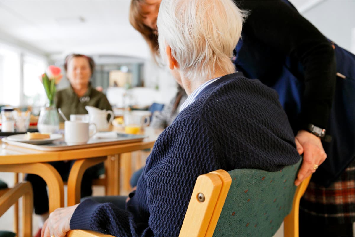 An older person is sitting at a table with other people in a bright room. A person standing nearby has one hand on the back of the chair.
