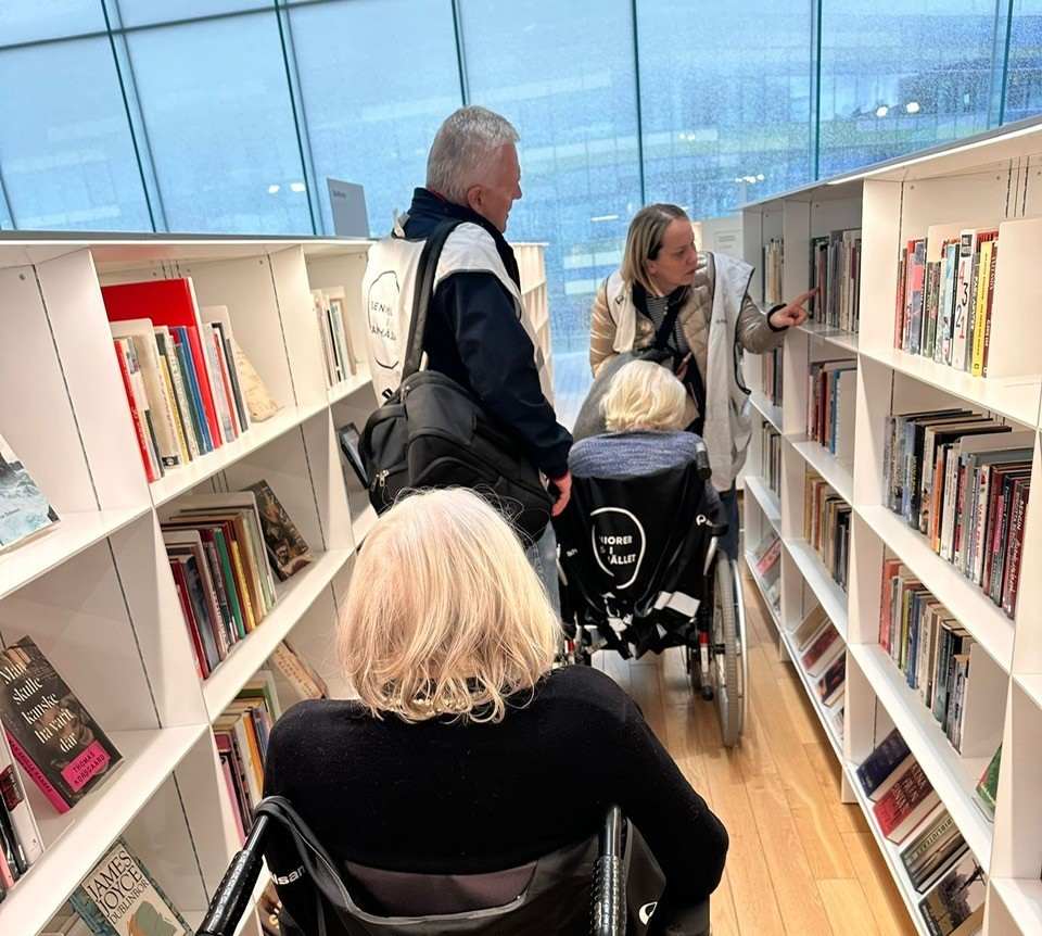 Två vårdare och två äldre i rullstol på biblioteket Ode bekantar sig med böcker i en hylla.