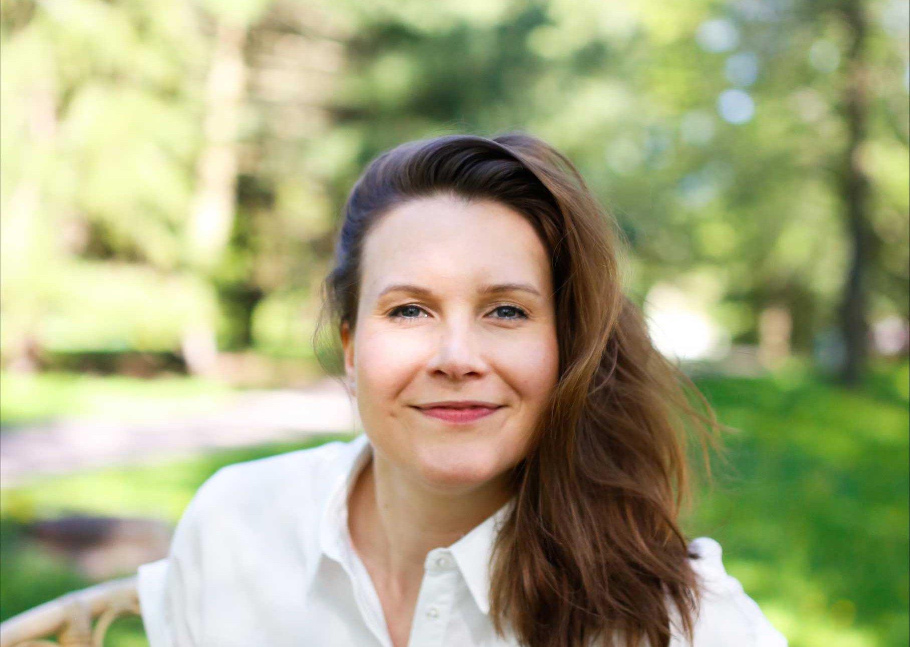 Porträtt av en leende kvinna iklädd en vit skjorta, i en grönskande utomhusmiljö.